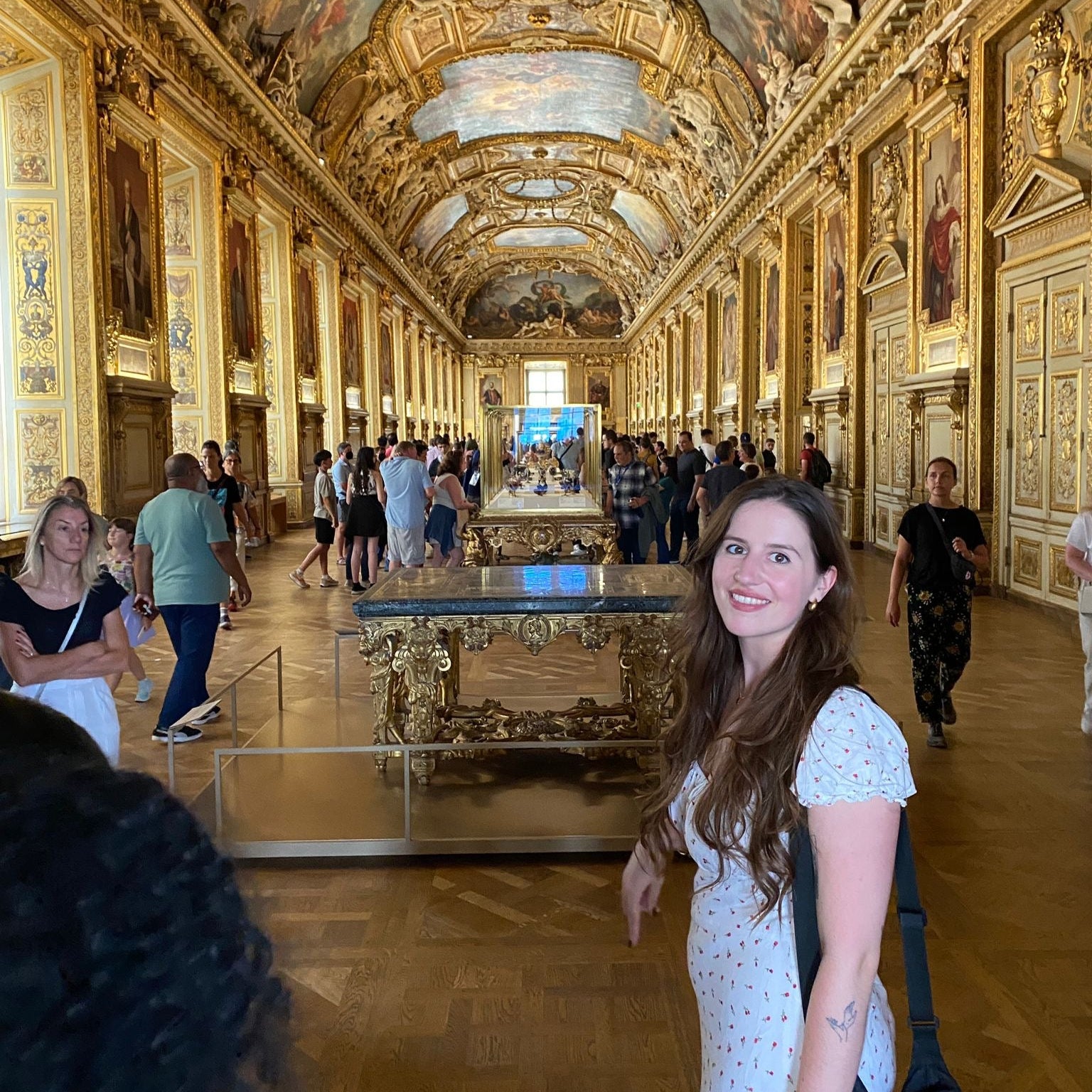 Woman standing in a grand hall of louvre with ornate gold ceiling and decor
